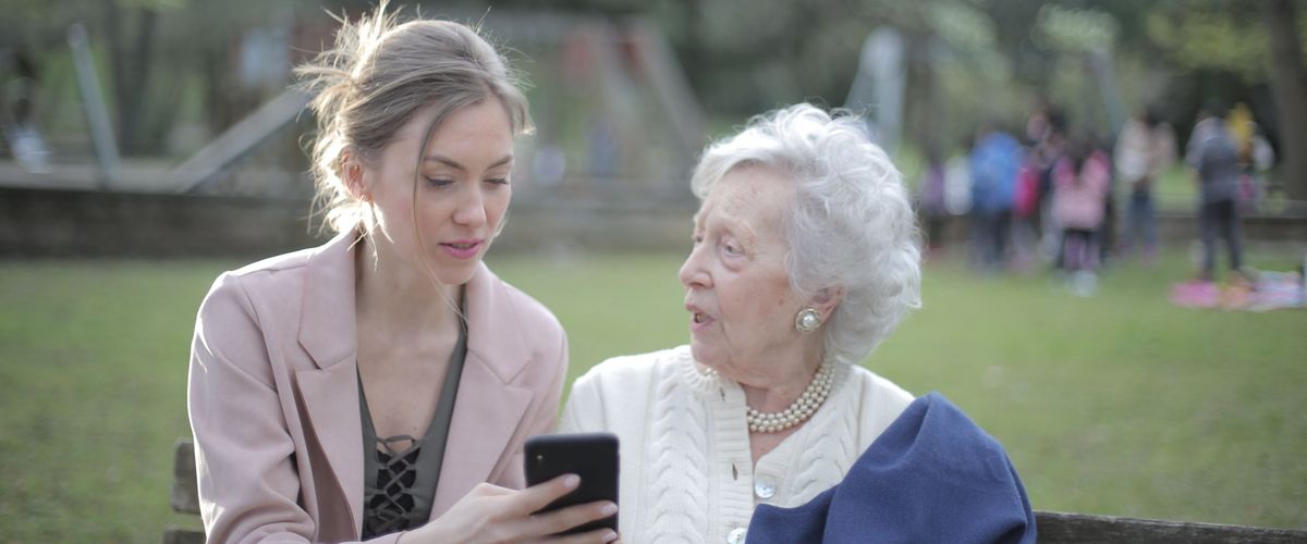 Mladá žena ukazuje starší paní na lavičce v parku chytrý telefon